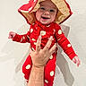adult_hand, baby, child, closeup, costume, cute, fashion, happy, held, indoor, infant, joyful, photography, polka_dots, portrait, red_outfit, shadow, smiling, sun_hat, wall