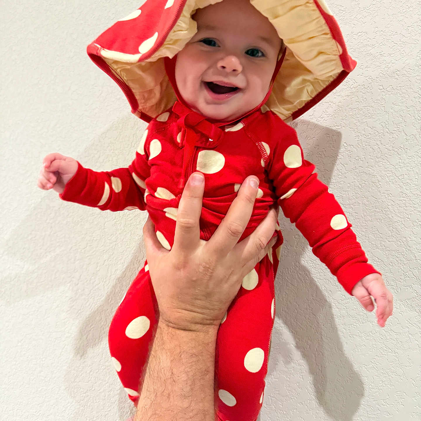 Tomas Matteo is registered to the contest to win money with this photo: baby, infant, child, smiling, happy, portrait, red_outfit, polka_dots, sun_hat, costume, held, adult_hand, indoor, wall, shadow, closeup, fashion, joyful, photography, cute