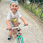 Rafaël a rejoint le concours — aidez-le/la à gagner de superbes lots ! toddler, child, smiling, bike, balance_bike, teddy_bear, cap, white_tshirt, shorts, black_shoes, pathway, iron_fence, trees, greenery, outdoor, daylight, happy, play, toy, nature