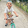 toddler, child, smiling, bike, balance_bike, teddy_bear, cap, white_tshirt, shorts, black_shoes, pathway, iron_fence, trees, greenery, outdoor, daylight, happy, play, toy, nature
