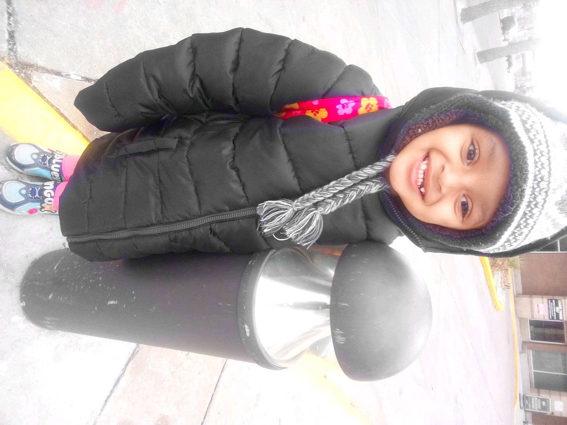 Bella is registered to the contest to win money with this photo: bollard, braided_tassel, child, coat_zipper, cold_weather, floral_backpack, happy_face, hat_pattern, knit_hat, outdoor, parking_area, pavement, sidewalk, smiling, sneakers, standing, street_curb, toddler, urban, winter_coat