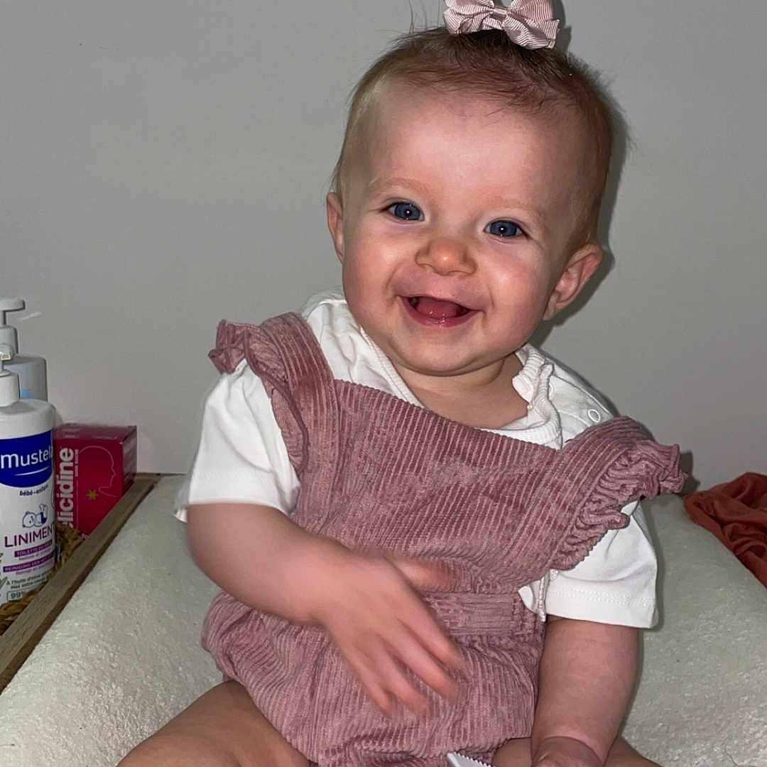 Elaïa a rejoint le concours — aidez-le/la à gagner de superbes lots ! baby, baby_care, blue_eyes, child, clothing, cute, face, hand, happy, indoor, infant, person, pink_bow, romper, room, sitting, skin, smiling, soft_surface, toy_knife