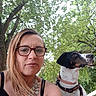 woman, glasses, dog, outdoor, tree, leaves, necklace, black_and_white_dog, harness, pet, nature, park, daylight, portrait, casual, person, animal, summer, greenery, selfie