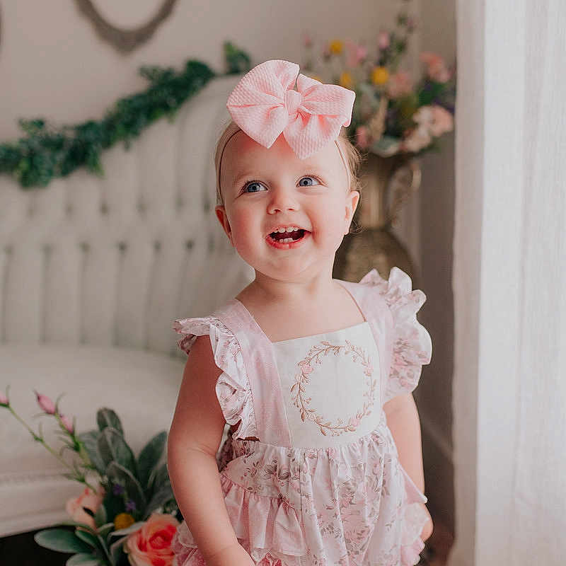 Kenzleigh is registered to the contest to win money with this photo: baby_toddler_clothing, child, clothing, dress, fashion_accessory, hair_accessory, head, headband, headgear, headpiece, iris, lace, peach, person, pink, ruffle, smile, standing, textile, toddler