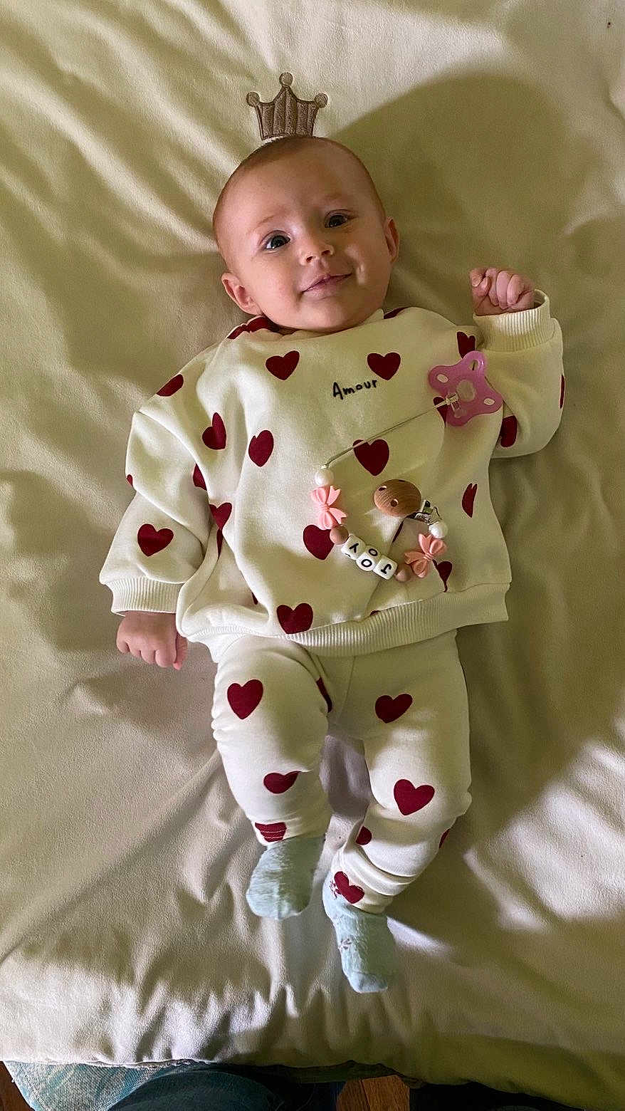 Joy participe au concours pour gagner de l'argent avec cette photo : baby, infant, heart_pattern, crown_illusion, pacifier, smiling, lying_down, clothing, socks, cute, soft_surface, indoors, child, person, portrait, happy, toy, warm_lighting, cozy, baby_girl