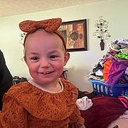 Kasey is registered to the contest to win money with this photo: child, toddler, smiling, headband, bow, clothing, laundry_basket, pile_of_clothes, portrait, indoor, home_interior, wall_art, man_partial, photobomb, rosy_cheeks, blanket, bed, cute, person, smile