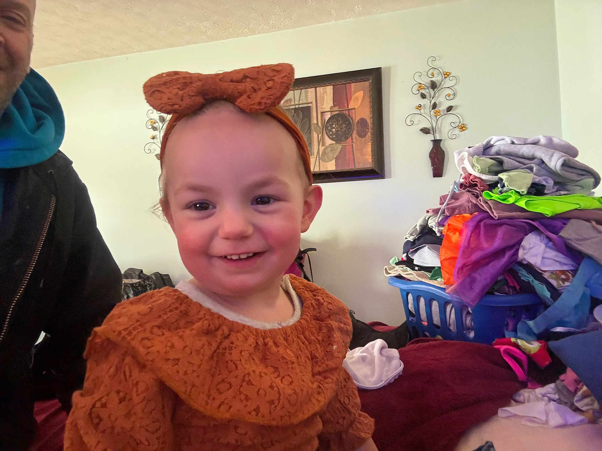 Kasey is registered to the contest to win money with this photo: child, toddler, smiling, headband, bow, clothing, laundry_basket, pile_of_clothes, portrait, indoor, home_interior, wall_art, man_partial, photobomb, rosy_cheeks, blanket, bed, cute, person, smile