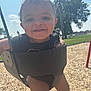 baby, child, swing, playground, smiling, happy, outdoors, sunny, grass, wood_chips, tree, blue_sky, dimples, drool, black_clothing, barefoot, metal_chain, infant, fun, park