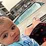 Jakai is registered to the contest to win money with this photo: baby, infant, child, poolside, swimming_pool, person, legs, shorts, cherry_pattern, outdoor, daylight, relaxation, apartment_building, concrete, water, face, curious, casual_clothing, lounge_chair, skin