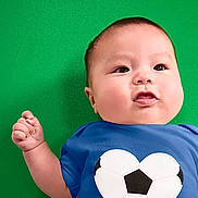 Ace is registered to the contest to win money with this photo: baby, infant, child, portrait, face, smiling, happy, blue_shirt, soccer_ball, raised_fist, green_background, indoor, close_up, sitting, round_face, short_hair, eyes, mouth, colorful, photography