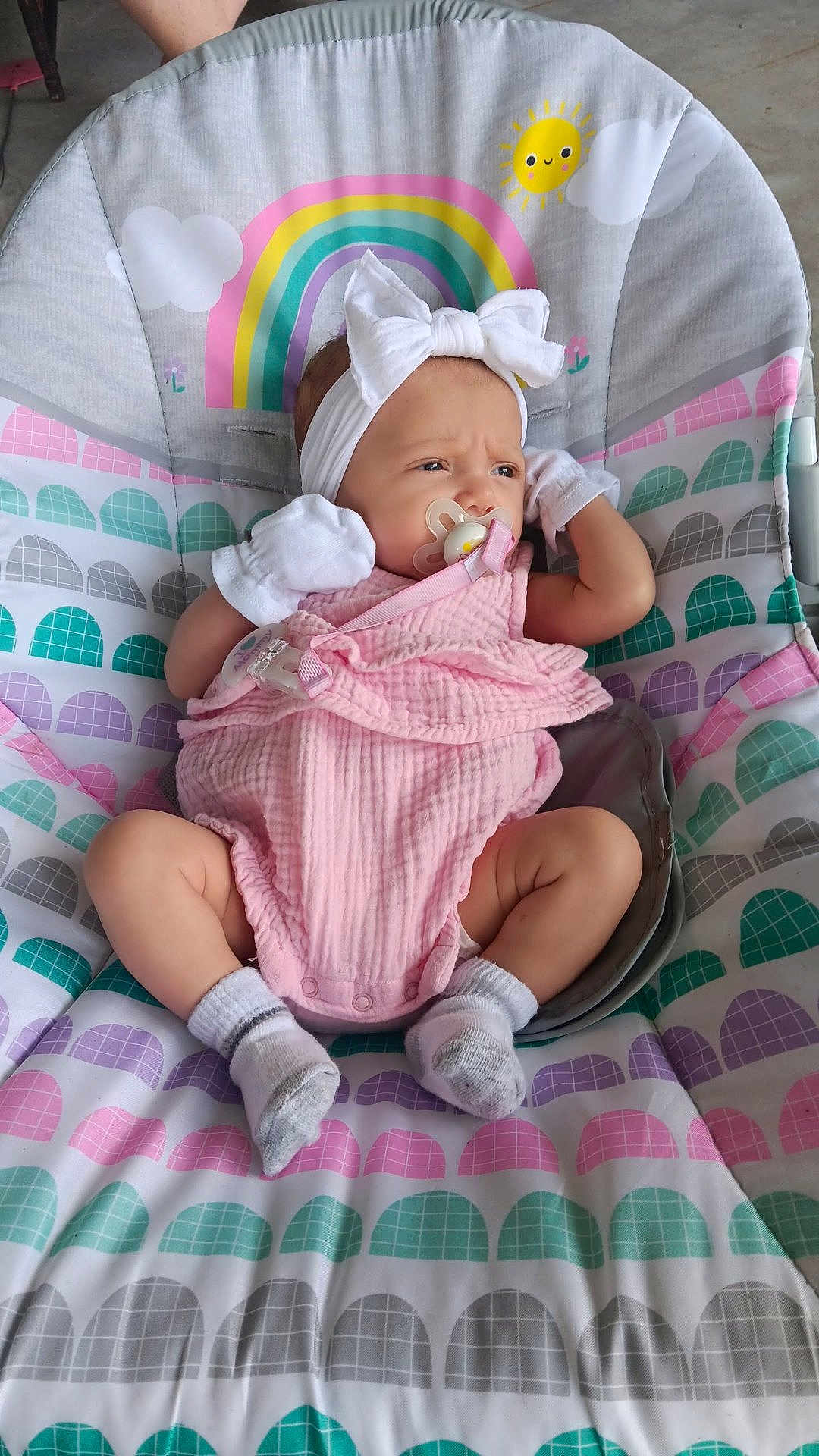 Kai is registered to the contest to win money with this photo: baby, infant, pink_outfit, white_bow, pacifier, baby_chair, rainbow_pattern, mittens, socks, cute, indoors, colorful, child, seat, newborn, headband, smiling_sun, clouds, patterned_fabric, relaxed