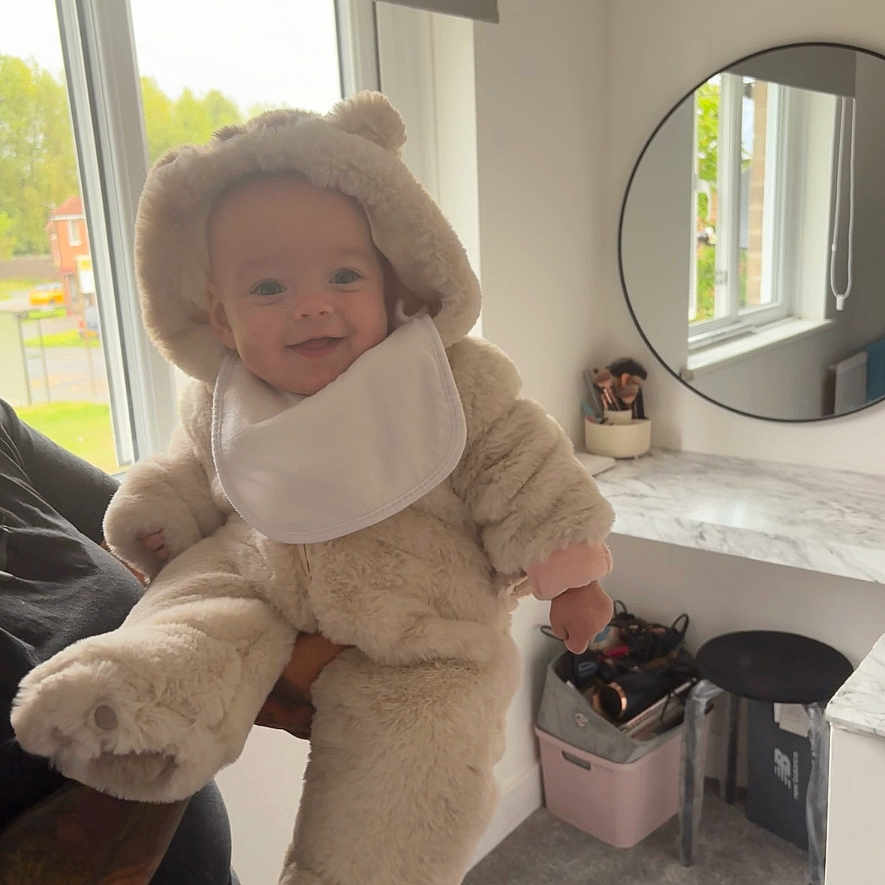 Harlie-Rose is registered to the contest to win money with this photo: adult_arm, baby, bear_costume, carpet, child, cozy, cute, fluffy_suit, holding, indoor, makeup_brushes, mirror, natural_light, portrait, smiling, stool, tattoo, vanity_table, white_wall, window