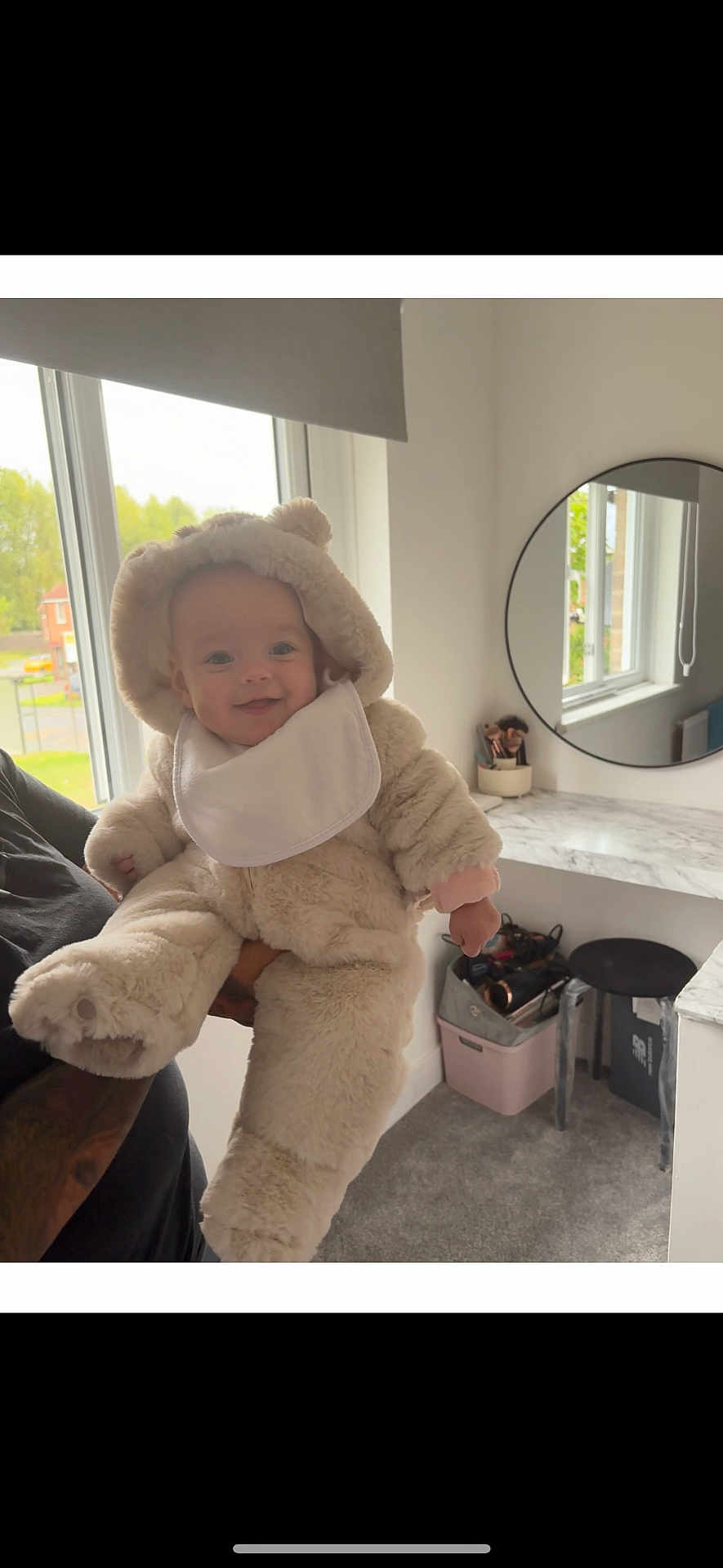 Harlie-Rose is registered to the contest to win money with this photo: baby, child, fluffy_suit, bear_costume, smiling, window, indoor, mirror, adult_arm, tattoo, carpet, vanity_table, makeup_brushes, stool, natural_light, white_wall, cozy, cute, holding, portrait