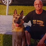 Rex a rejoint le concours — aidez-le/la à gagner de superbes lots ! dog, german_shepherd, man, person, outdoor, grass, parked_car, vehicle, signpost, sitting, portrait, companion, pet, leash, collar, tongue_out, upright_ears, glasses, black_shirt, daylight