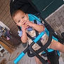 toddler, child, stroller, snack, porch, wooden_floor, smiling, blue, clothing, seatbelt, outdoor, baby, person, hand, foot, door, window, rope, chair, house