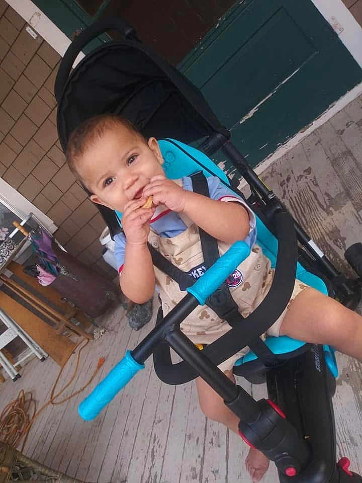 Mateo is registered to the contest to win money with this photo: toddler, child, stroller, snack, porch, wooden_floor, smiling, blue, clothing, seatbelt, outdoor, baby, person, hand, foot, door, window, rope, chair, house