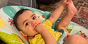Adler is registered to the contest to win money with this photo: baby, infant, child, yellow_shirt, baby_seat, hand_in_mouth, foot_in_air, indoor, carpet, furniture, curly_hair, playful, cute, seatbelt, colorful, patterned_rug, comfortable, happy, young_child, portrait