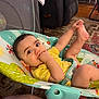baby, infant, child, yellow_shirt, baby_seat, hand_in_mouth, foot_in_air, indoor, carpet, furniture, curly_hair, playful, cute, seatbelt, colorful, patterned_rug, comfortable, happy, young_child, portrait