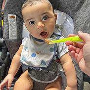 Adler is registered to the contest to win money with this photo: baby, high_chair, bib, baby_food, spoon, hand, feeding, indoor, child, cute, expression, person, seat, clothing, food, table, infant, mouth_open, looking, skin