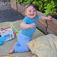 Samuel a rejoint le concours — aidez-le/la à gagner de superbes lots ! baby, child, smiling, outdoor, toy, mat, cushion, plant, greenery, wooden_planter, grass, happy, clothing, blue_shirt, pants, face, person, play, nature, daylight