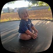 Brahim is registered to the contest to win money with this photo: baby, child, smiling, trampoline, outdoor, sunlight, grass, fence, farm, diaper, blue_shirt, happy, sitting, nature, shadow, person, cute, play, infant, tooth