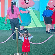 Prisha is registered to the contest to win money with this photo: child, girl, toddler, portrait, standing, red_dress, sunglasses, smiling, outdoor, theme_park, peppa_pig, mascot, cartoon, playground, pink, velvet_rope, artificial_grass, adults, colorful, family_outing
