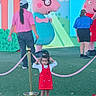 child, girl, toddler, portrait, standing, red_dress, sunglasses, smiling, outdoor, theme_park, peppa_pig, mascot, cartoon, playground, pink, velvet_rope, artificial_grass, adults, colorful, family_outing