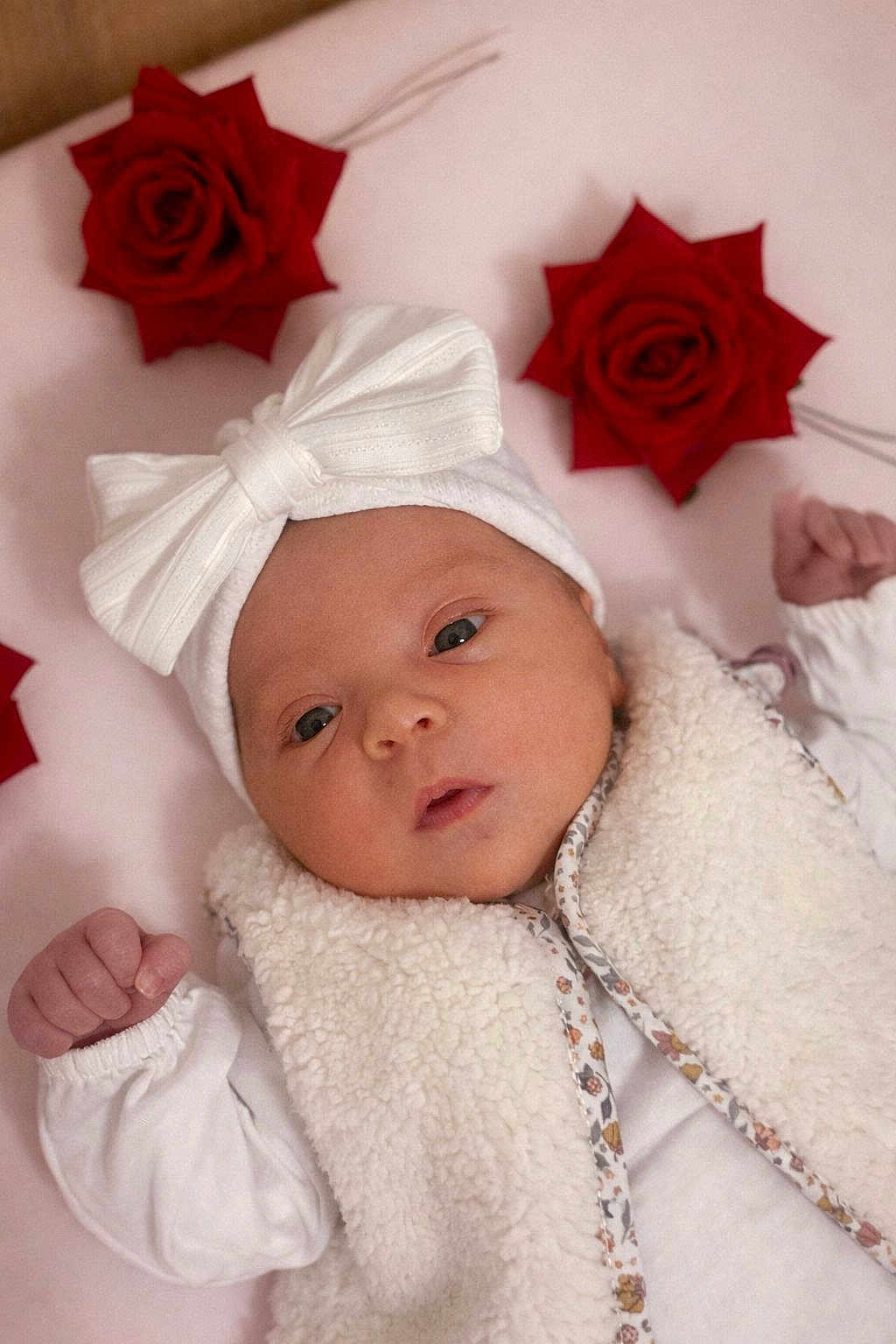 Emma participe au concours pour gagner de l'argent avec cette photo : baby, infant, white_bow, fluffy_vest, red_roses, pink_background, cute, newborn, portrait, face, headwear, soft_texture, small_fists, lying_down, closeup, adorable, baby_clothing, decorative_flowers, innocent, expression