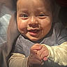 adorable, baby, child, clapping, close_up, clothing, eyes, face, gray_shirt, hands, happy, indoors, infant, long_sleeves, photograph, portrait, seated, smiling, soft_background, warm_lighting