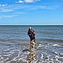 Ezra is registered to the contest to win money with this photo: person, dog, water, ocean, waves, sky, clouds, beach, leash, casual_clothing, cap, happy, outdoor, summer, walking, pet, sea, reflection, fun, sunlight