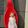 child, girl, dress, red_spoon, sculpture, peace_sign, smile, outdoor, sunlight, shadow, rocks, concrete_wall, happy, blonde_hair, casual_shoes, playful, posing, young, cute, colorful