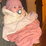 Jade participe au concours pour gagner de l'argent avec cette photo : baby, pacifier, blanket, knitted_hat, adult, green_hoodie, cozy, wrapped, warm_clothing, indoor, portrait, child, person, face, holding, soft_texture, pink_blanket, curious_eyes, night, warm_lighting