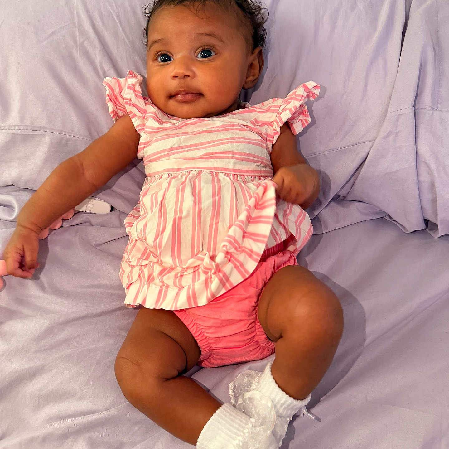 Saige is registered to the contest to win money with this photo: baby, infant, child, pink_clothing, striped_shirt, shorts, white_socks, bedspread, lavender, smiling, cute, indoor, portrait, person, relaxing, lying_down, young_child, happy, soft_fabric, cozy