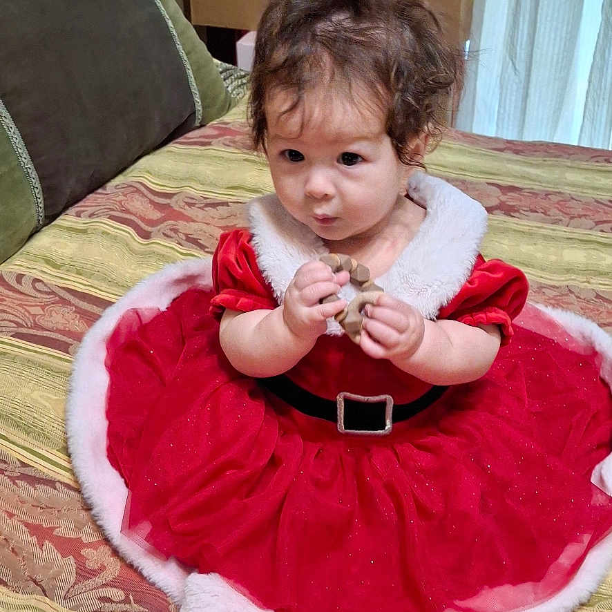 Myonie joined the competition — help win amazing prizes! baby, child, red_dress, holiday_clothing, bedspread, indoor, curly_hair, sitting, person, furniture, lamp, cardboard_box, curtain, patterned_fabric, holding_object, cute, young_child, home, festive, portrait