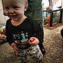 child, baby, toddler, smile, face, standing, birthday_shirt, clothing, patterned_pants, toy, carpet, indoor, home, playroom, adult_legs, hand, candid, closeup, happy, floor