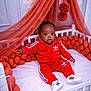 baby, child, crib, red_clothing, tracksuit, adidas, sneakers, orange_canopy, orange_cushions, indoor, portrait, cute, infant, sitting, white_furniture, plush, curly_hair, looking_at_camera, cozy, home