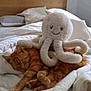 cat, orange_tabby, sleeping, plush_octopus, stuffed_animal, bed, white_comforter, pillow, cozy, cute, indoor, nap, soft_toy, tentacles, resting, feline, pet, home, relaxation, sleep