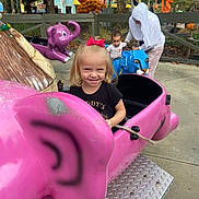 Cheryl is registered to the contest to win money with this photo: child, girl, toddler, adult, amusement_park, ride, pink_elephant, blue_animal_ride, smile, outdoor, playground, person, tree, fence, concrete, clothing, bow, fun, daytime, family