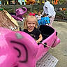 child, girl, toddler, adult, amusement_park, ride, pink_elephant, blue_animal_ride, smile, outdoor, playground, person, tree, fence, concrete, clothing, bow, fun, daytime, family