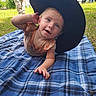baby, child, blanket, hat, outdoor, grass, person, plaid, blue, curious, joyful, headwear, clothing, vehicle, trailer, nature, sunlight, smile, portrait, recreation