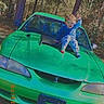 Damon joined the competition — help win amazing prizes! child, car, green_car, outdoor, trees, boy, hood, waving, smiling, daylight, nature, grass, casual_clothing, automobile, parked_car, child_playing, young_boy, vehicle, sunlight, happy
