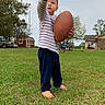 Damon is registered to the contest to win money with this photo: child, toddler, football, grass, barefoot, pointing, outdoor, sky, tree, house, lawn, casual_clothing, young_child, daytime, person, play, sport, nature, residential_area, cute