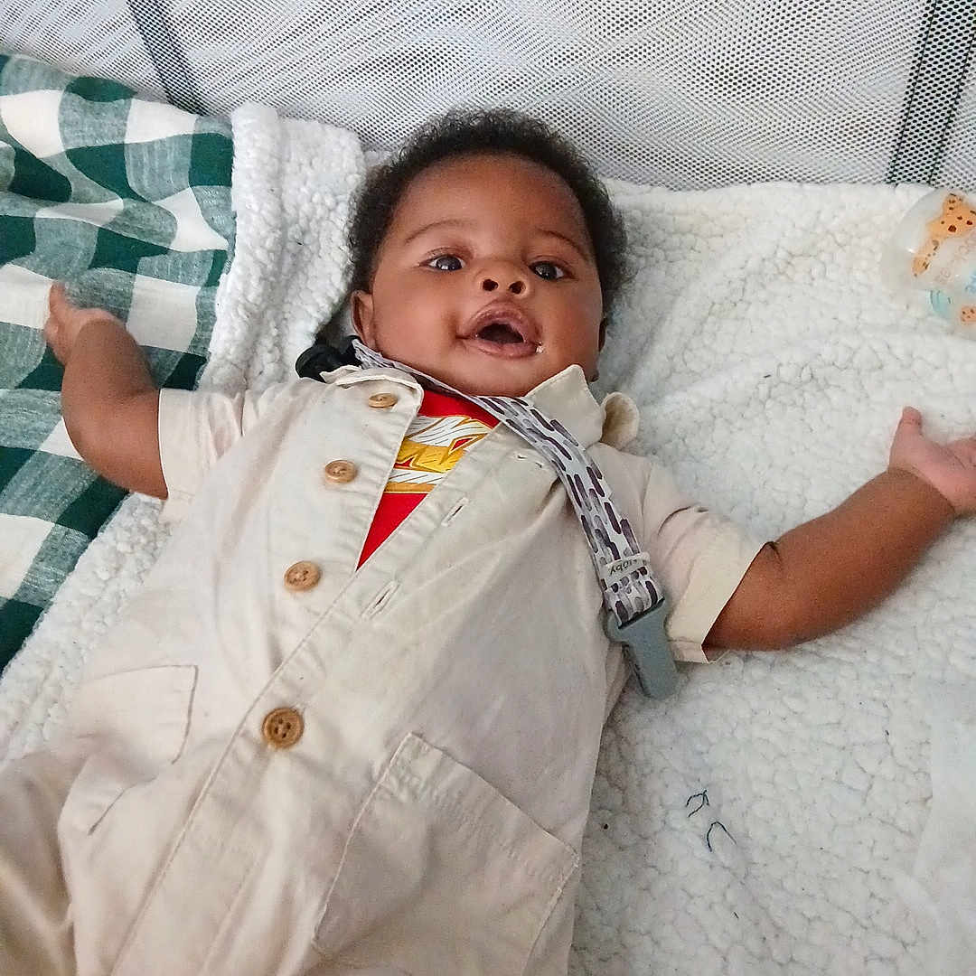 A'Maris is registered to the contest to win money with this photo: arm, baby, blanket, child, clothing, cozy, cute, expression, face, happy, indoor, infant, lying_down, pacifier_clip, person, playpen, smiling, soft_texture, toddler, wooden_buttons