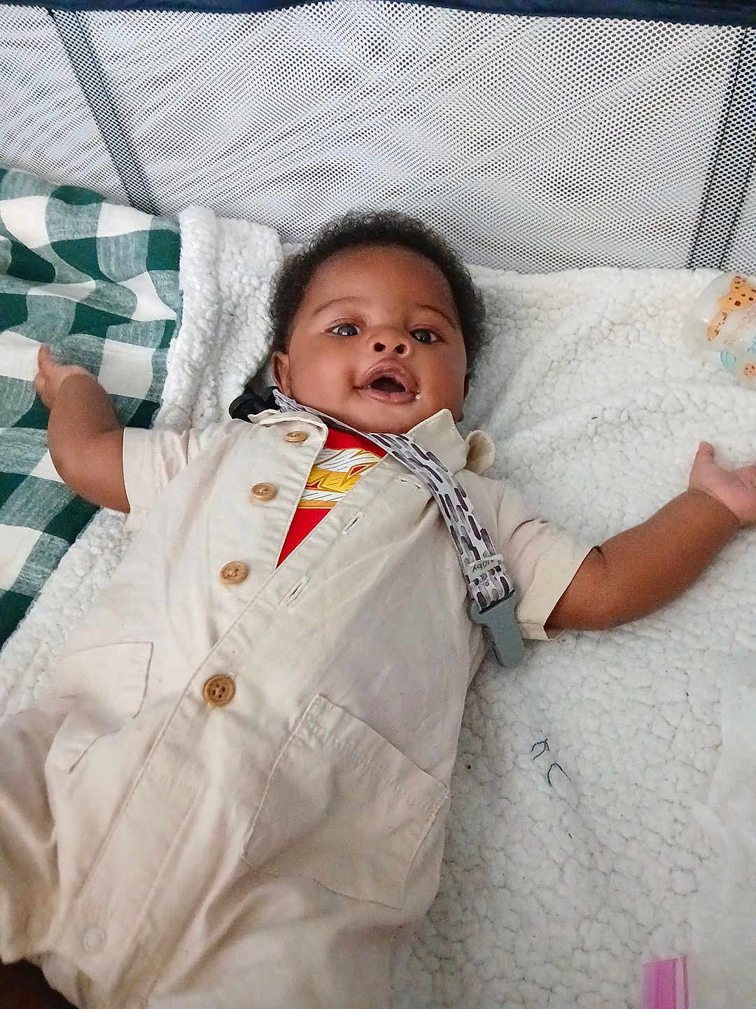 A'Maris is registered to the contest to win money with this photo: baby, infant, playpen, blanket, clothing, wooden_buttons, pacifier_clip, smiling, happy, lying_down, indoor, child, cute, cozy, soft_texture, face, person, expression, arm, toddler