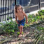 Ugo a rejoint le concours — aidez-le/la à gagner de superbes lots ! bare_chest, blue_shorts, bushes, child, curly_hair, daylight, fence, garden, greenery, leaves, nature, outdoor, person, plants, sandals, sidewalk, sunlight, toddler, walking, young_child