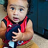 baby, child, curly_hair, slipper, carpet, red_shirt, tan_shorts, barefoot, indoors, wide_eyes, expression, holding, sitting, floor, storage_bin, cute, infant, person, casual_clothing, playful