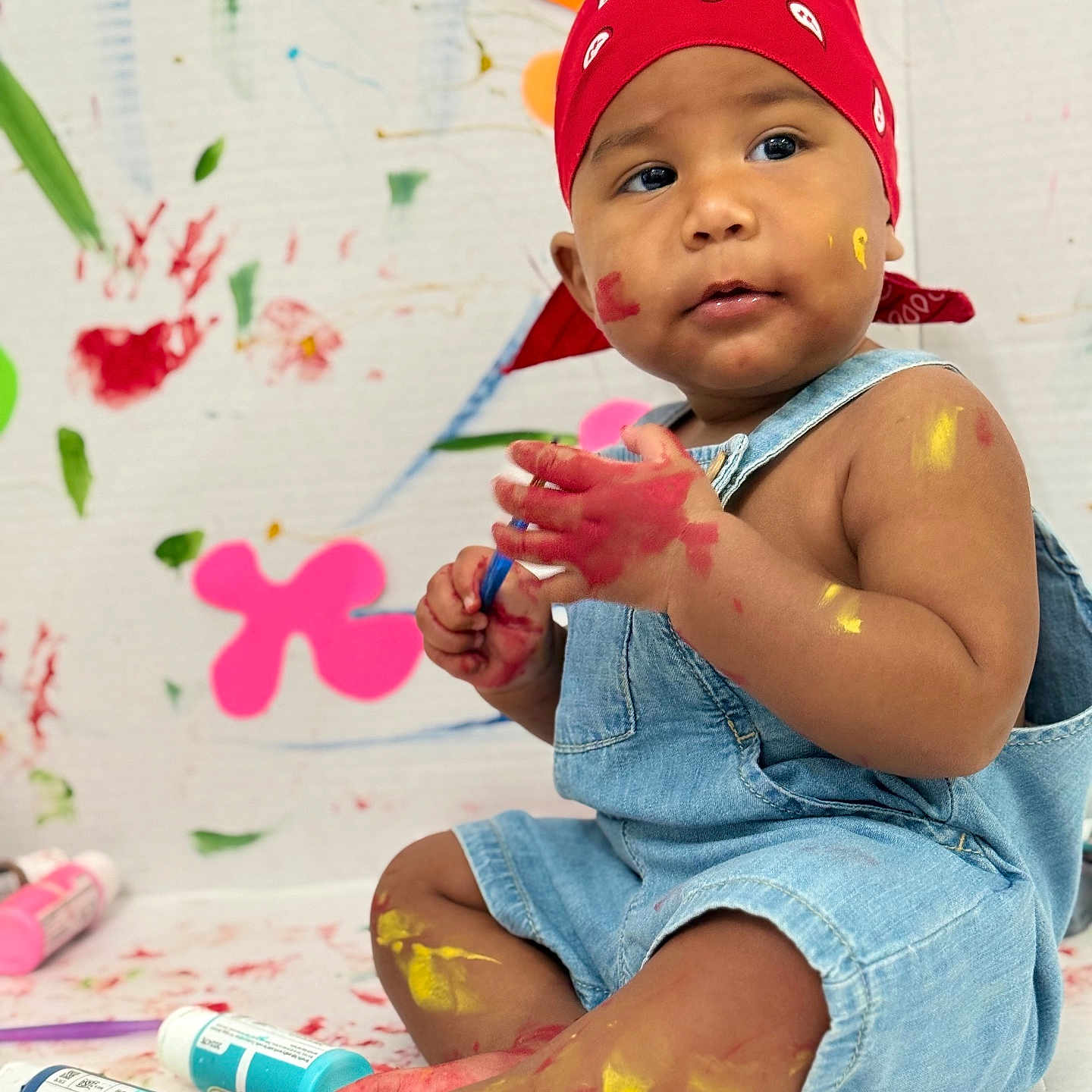Fred joined the competition — help win amazing prizes! toddler, child, paint, colorful, bandana, denim_overalls, sitting, face_paint, feet, hands, playful, art, messy, indoor, baby, expression, skin, background, floor, bottle