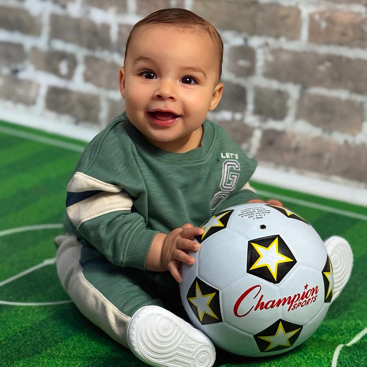 Zylan is registered to the contest to win money with this photo: baby, brick_wall, casual_clothing, child, cute, face, floor, green_carpet, hands, happy, indoor, playful, portrait, sitting, smiling, soccer_ball, sports, toy, white_shoes, young_child