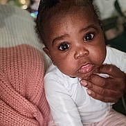 Calyah is registered to the contest to win money with this photo: baby, blanket, child, close_up, clothing, curious, cute, eyes, face, hand, indoors, infant, knit_blanket, person, pink_pants, portrait, seated, skin, soft_light, white_shirt