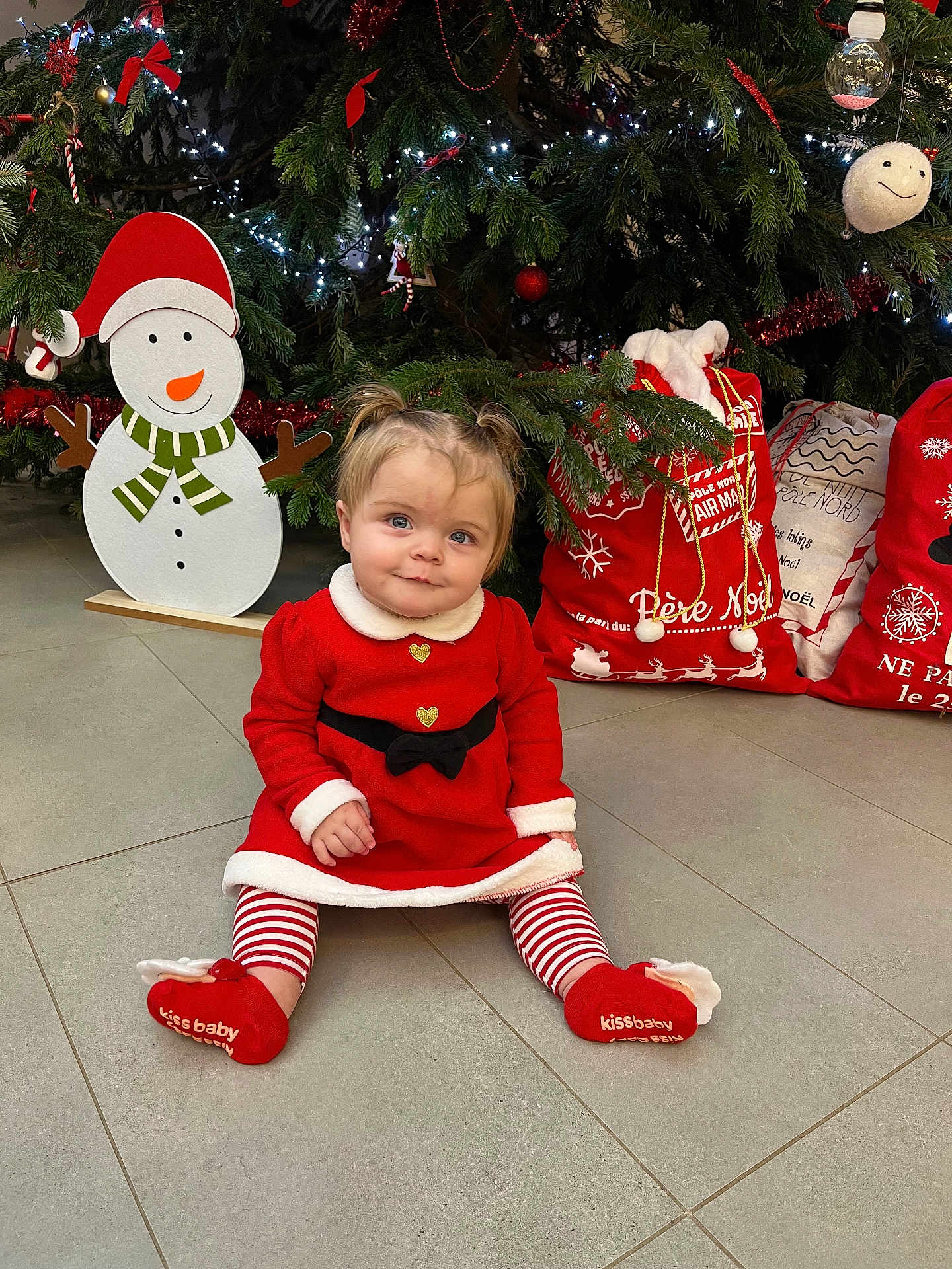 Marion a rejoint le concours — aidez-le/la à gagner de superbes lots ! baby, celebration, child, christmas_bags, christmas_tree, cute, face, festive, floor, holiday_decor, indoor, pants, person, pigtails, red_dress, seasonal, shoes, smiling, snowman_decoration, striped_leggings